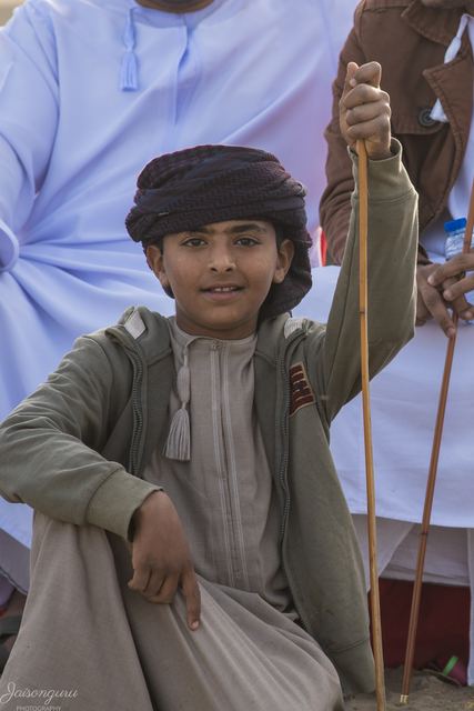 Young boy in a dishdasha and turban seated with a herding stick at the livestock market