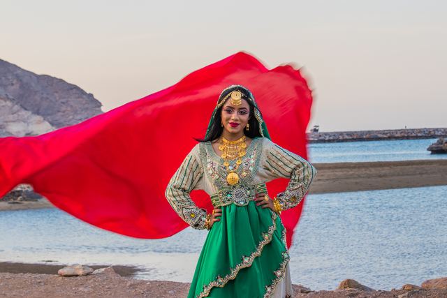 Woman in traditional dress on the shore, red headscarf lifting in the wind