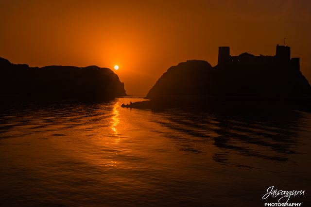 Deep orange sunrise between two headlands with a hilltop fort in silhouette