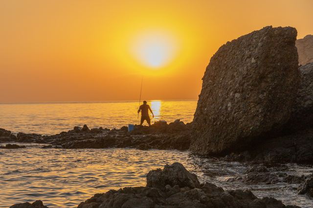 Fisherman casting from the rocks, silhouetted against a hazy sunrise